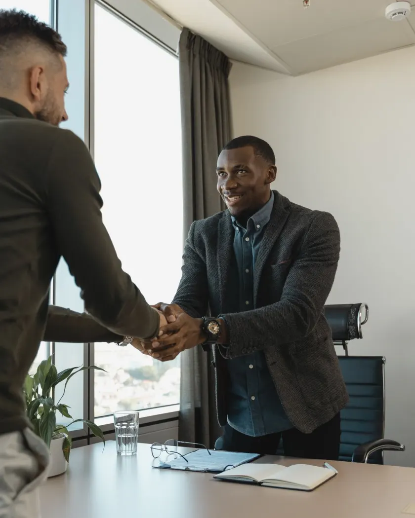 Two men shaking hands across a desk in a bright office, a notebook and glasses between them.