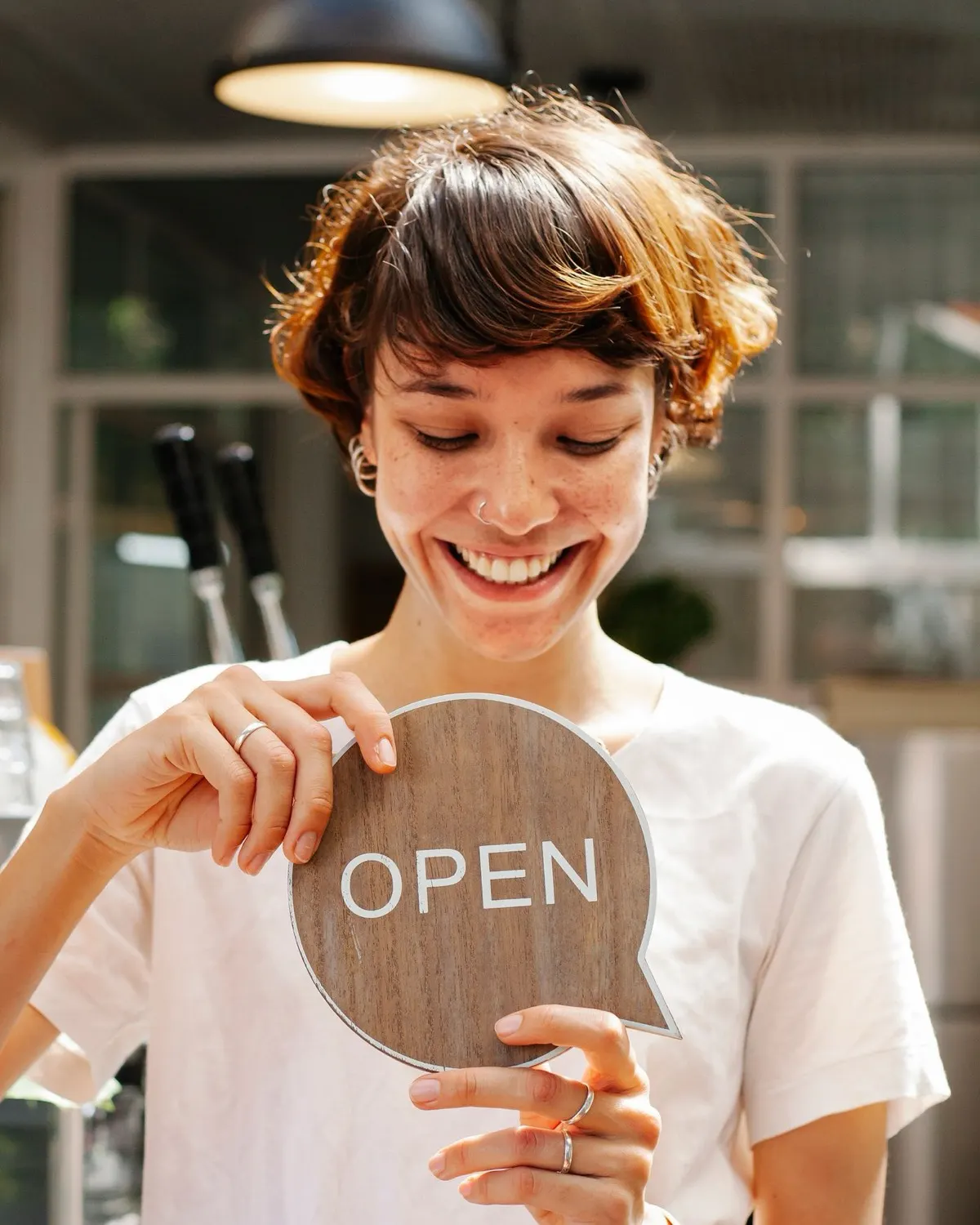 A café owner smiling as she holds up a wooden Open sign behind her counter.